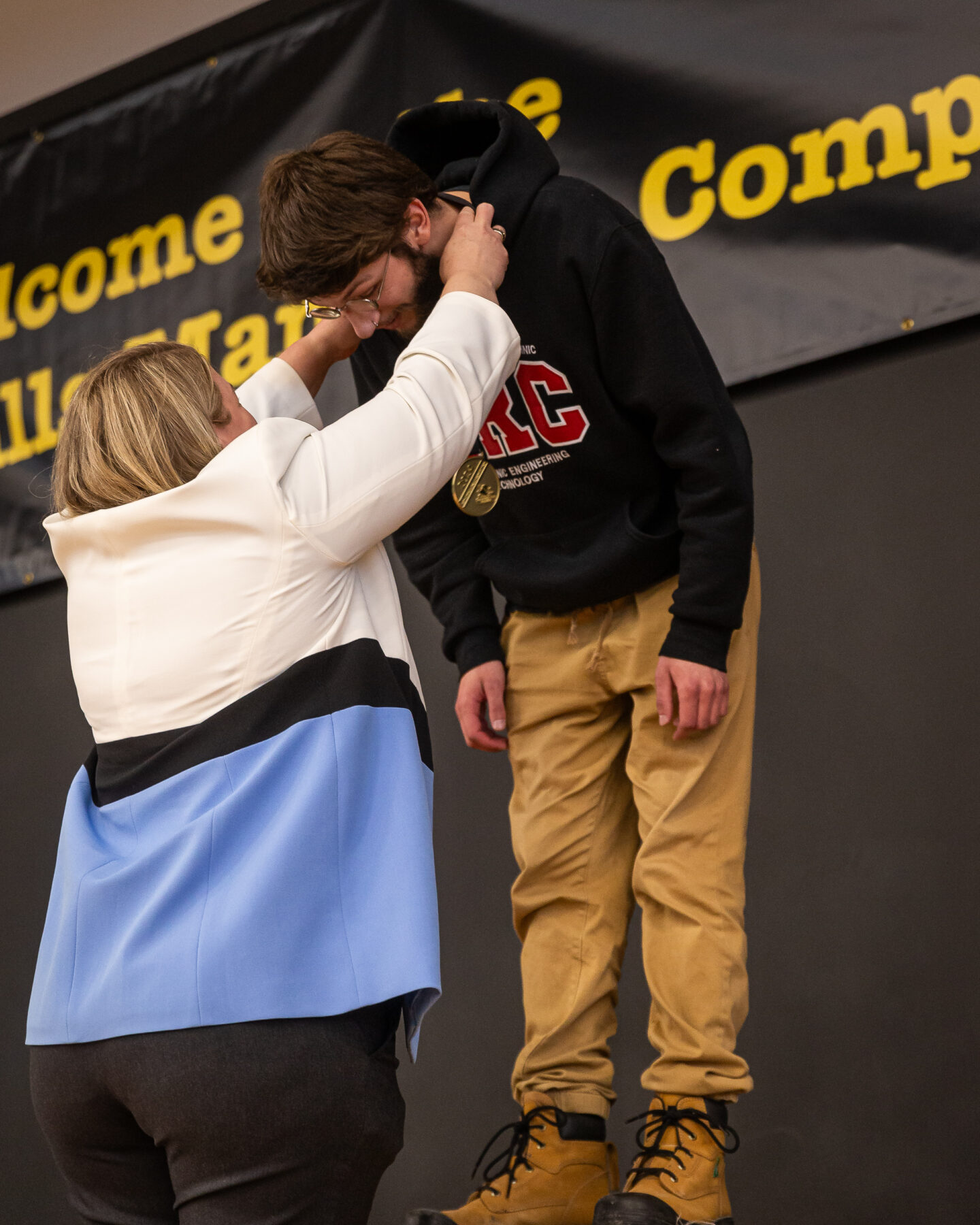 Student wearing an RRC Polytech sweater leans forward on a stage as a woman drapes a medal around the student's neck.