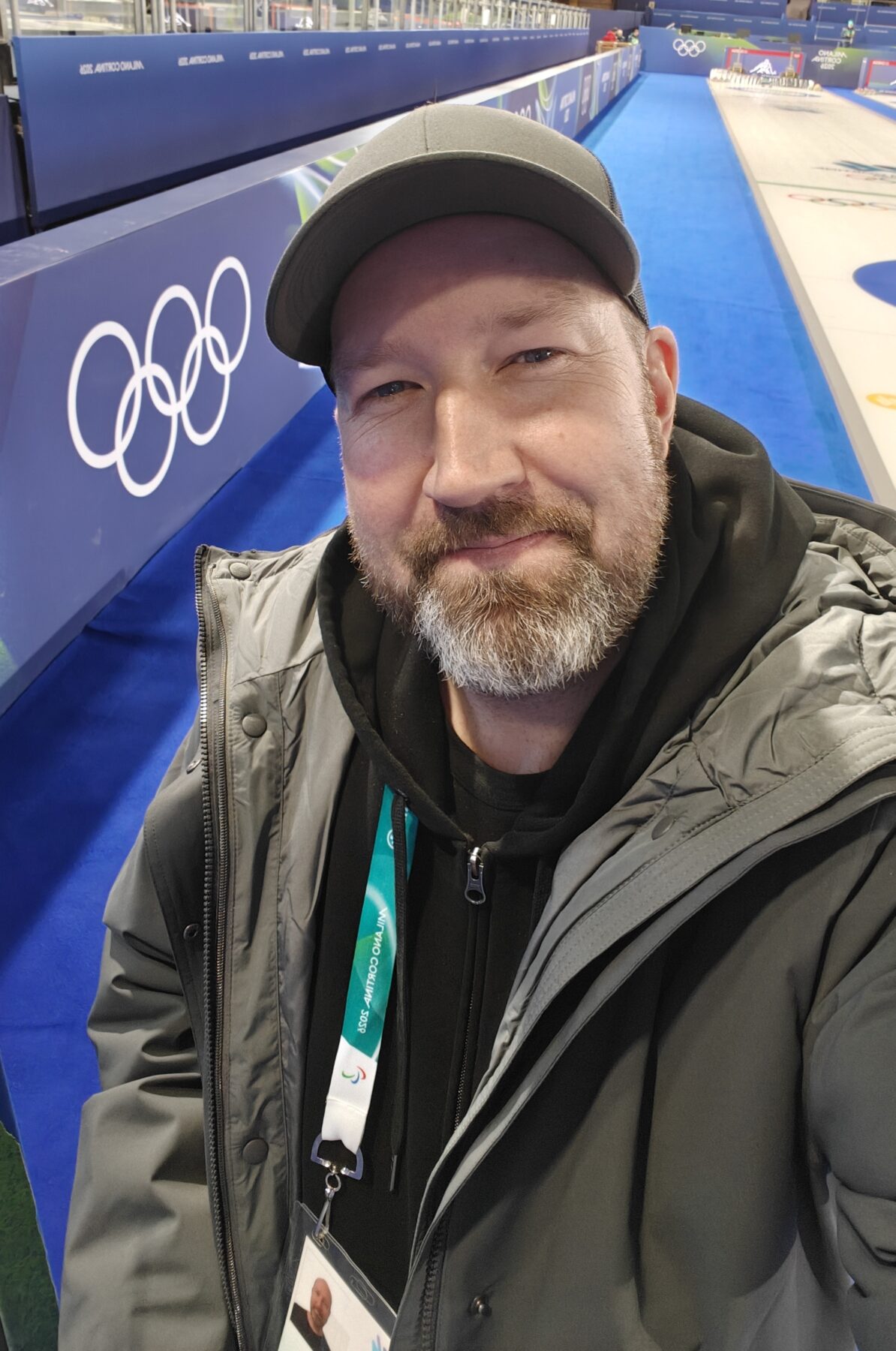 A person wearing a hat and parka stands in a curling rink and takes a selfie.