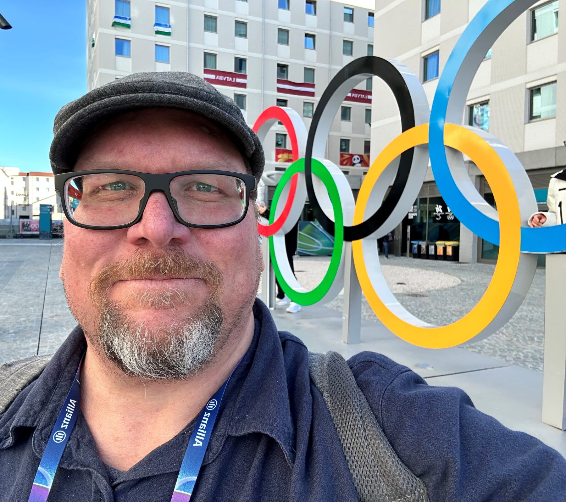A person wearing a hat and glasses takes a selfie in front of the Olympics rings.