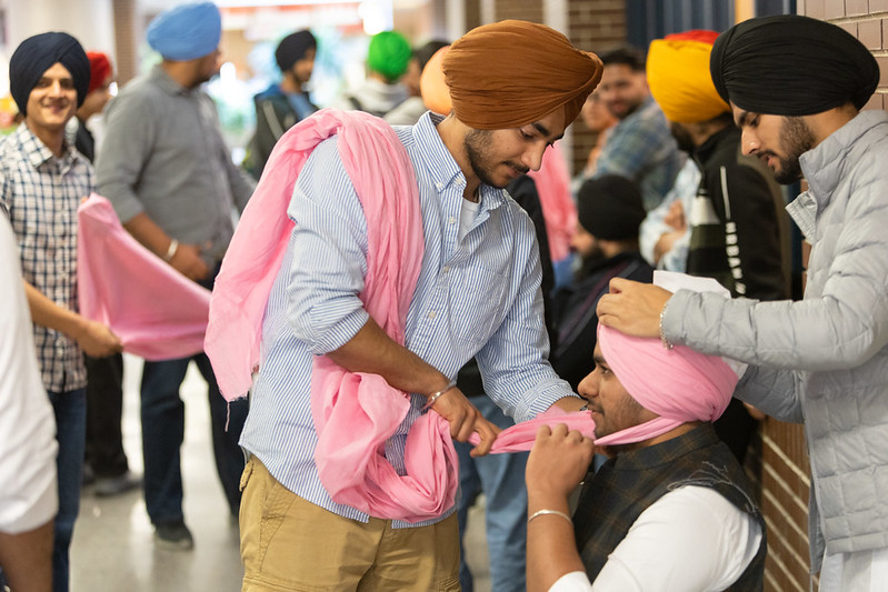 A person sits in a chair, while another person wraps a pink turban on their head,