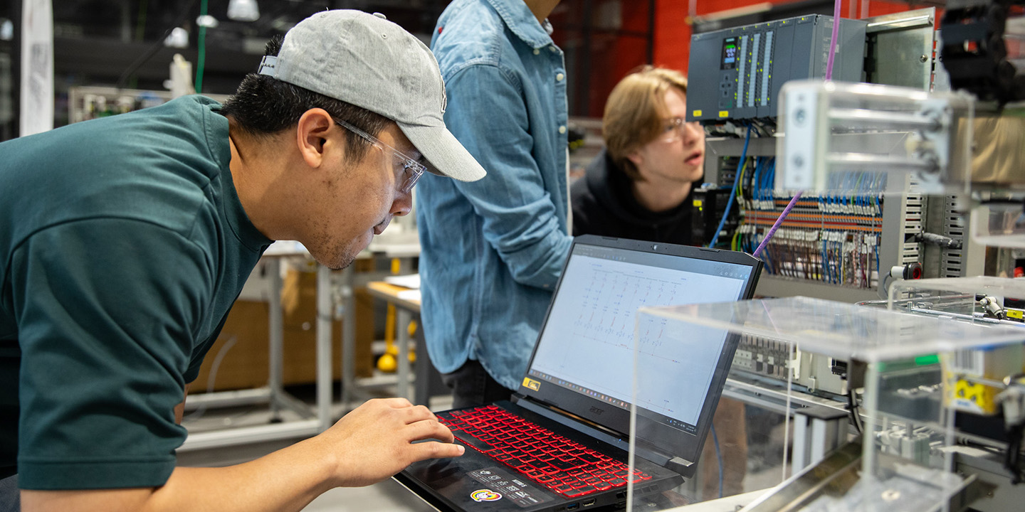 A student wearing a gray cap works on a laptop, in a lab setting.