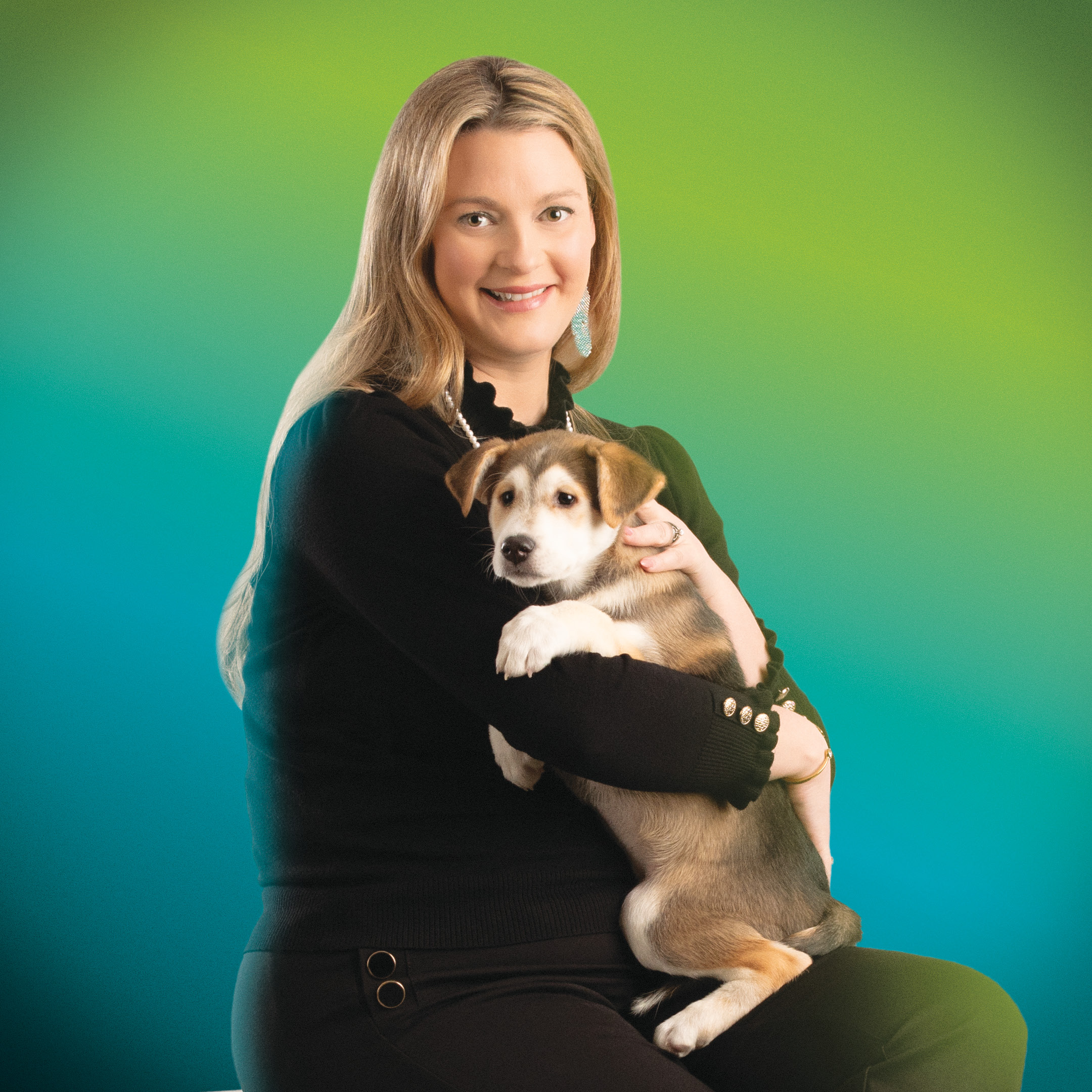 RRC Polytech graduate Jessica Miller is seated and wearing all black. She holds a brown and white puppy in her arms in front of a green and blue backdrop.