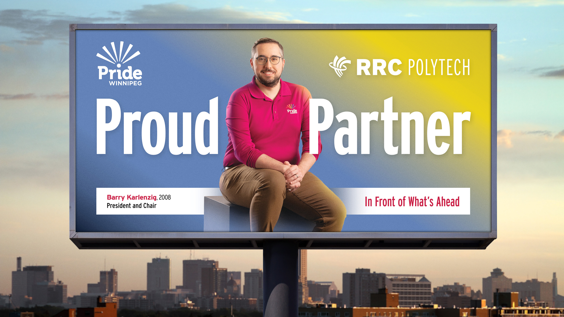 RRC Polytech graduate Barry Karlenzig wears a fuchsia Pride Winnipeg polo shirt and tan pants and is in a seated position on a billboard as part of the College's Polytechnic Proud campaign. The billboard rises above the Winnipeg city skyline.