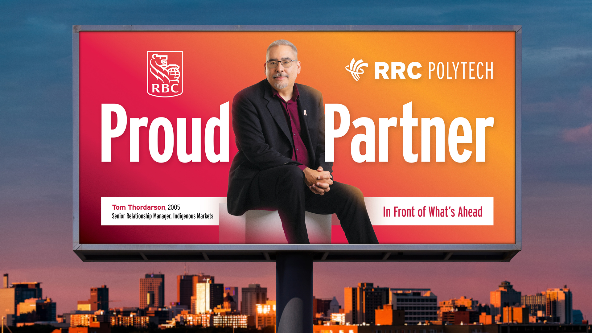 RRC Polytech graduate Tom Thordarson is featured on a billboard above the Winnipeg skyline. He is seated and wearing a black suit with dark red collared shirt. The billboard features the RBC logo and the RRC Polytech logo. The billboard is part of the College's Polytechnic Proud campaign, which featured 41 alumni across the province.