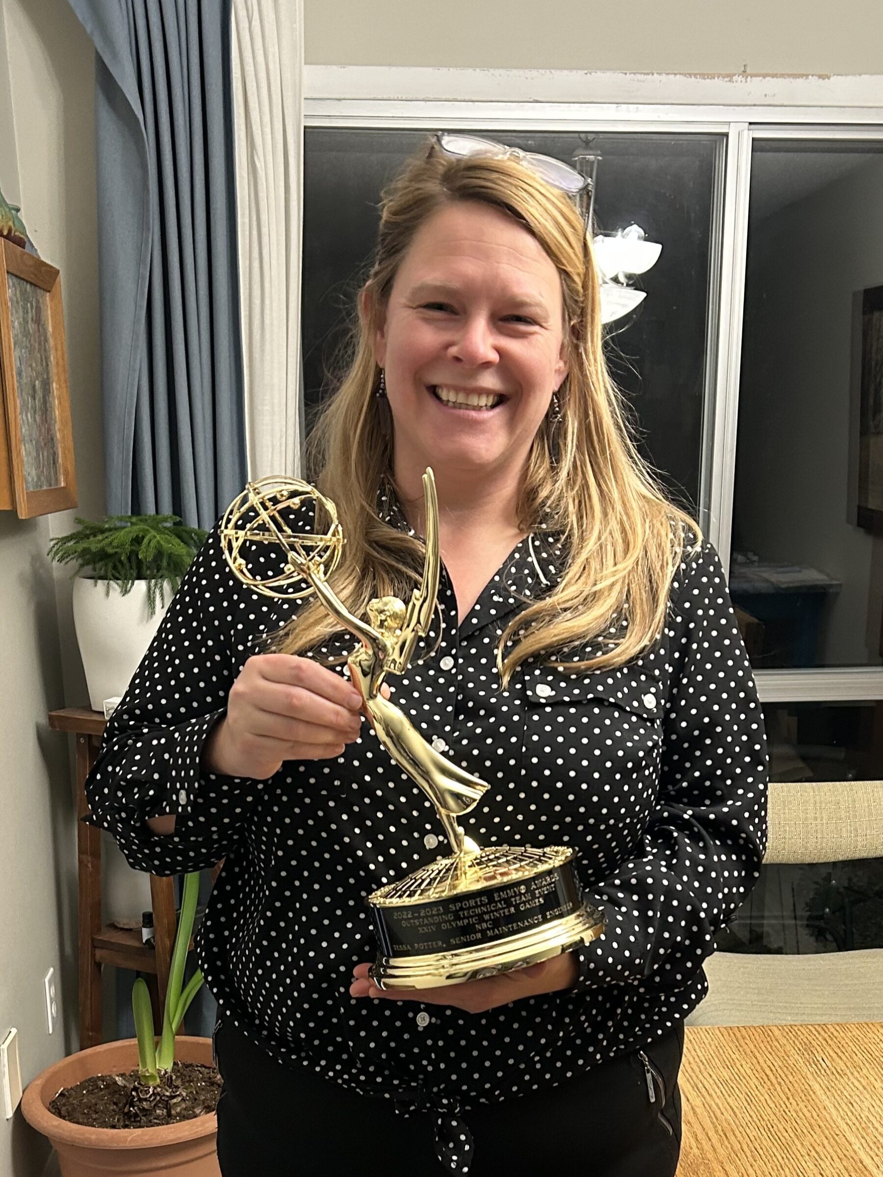 A woman with blonde hair wearing a black shirt and black pants stands in front of a window holding a golden figurine, an Emmy award.