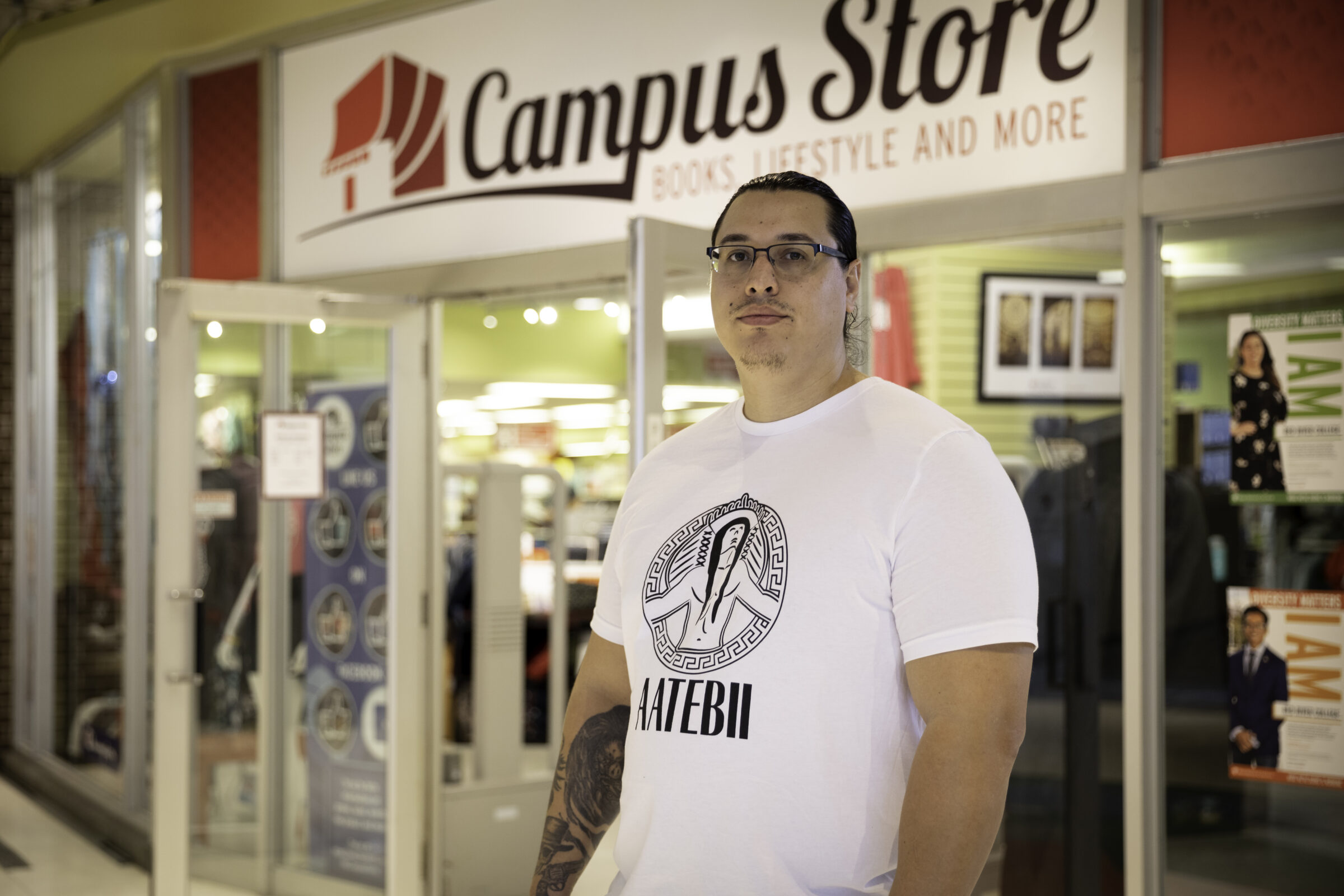 RRC Polytech graduate Sean Rayland-Boubar is wearing a white T-shirt and glasses and stands on the right-hand side of the Notre Dame Campus bookstore, beneath the Campus Store sign.