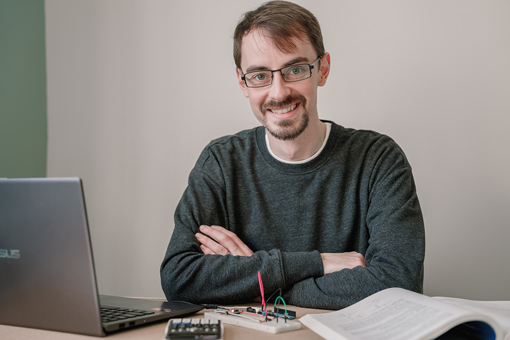 Stephen Klatt sitting at his desk at home. He has a laptop, calculator, a circuit board and a textbook in front of him.