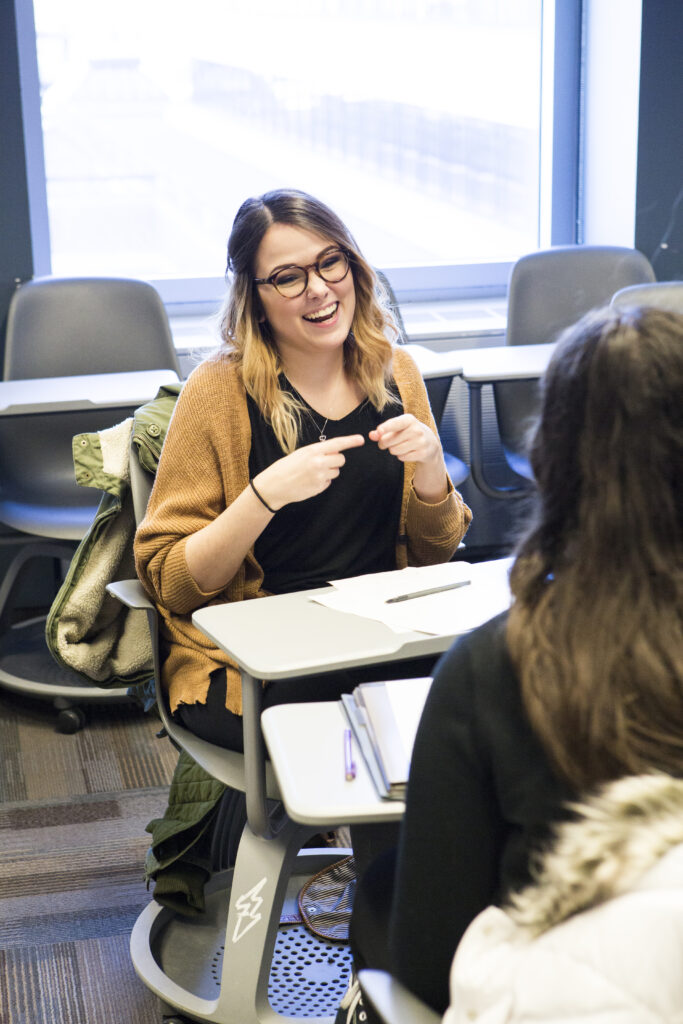 A student in ASL studies sitting in a classroom, communicating with another student using sign language.