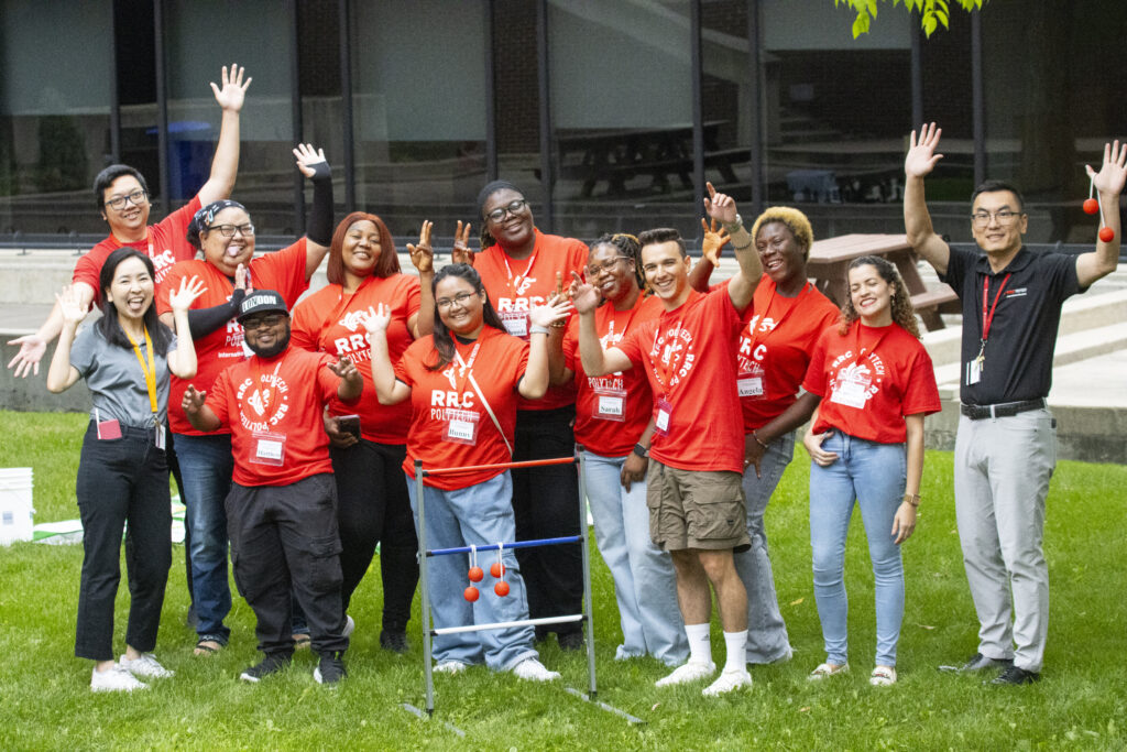 A group of smiling students and staff from RRC Polytech pose together outdoors outside Notre Dame Campus. Most are wearing red RRC Polytech T-shirts and name tags, with their hands raised in celebration. They appear happy and engaged during a campus event or activity.