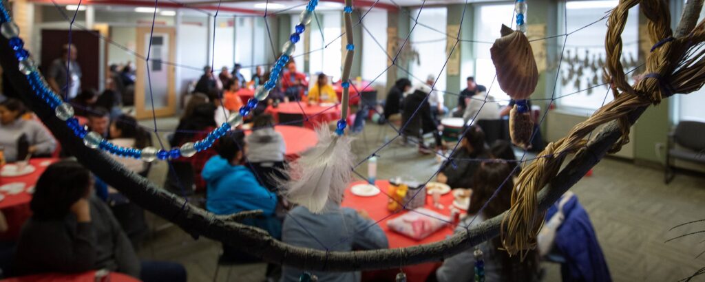 A gathering of people seated at round tables in the Indigenous Student Support Centre, viewed through a large dreamcatcher decorated with beads, feathers, and braided fibers.
