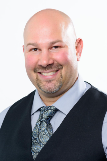 A person wearing a blue tie and black vest smiles at the camera.