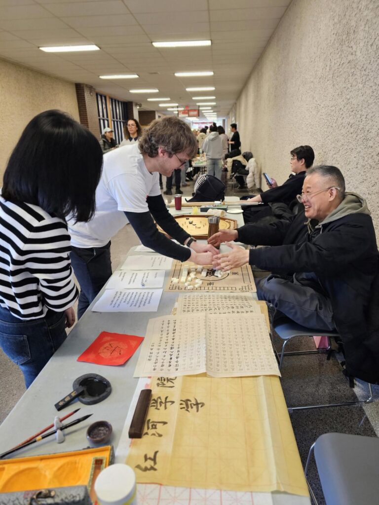People engaging in a tabletop game and calligraphy activity at a cultural booth in a campus hallway.