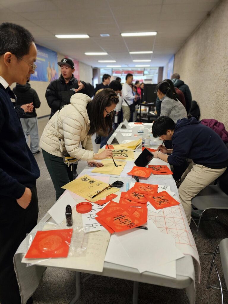 People creating calligraphy on red paper at a long activity table.