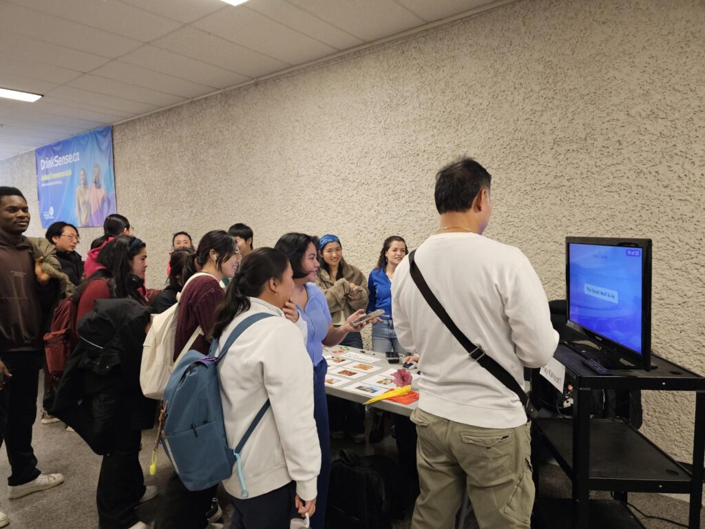 A crowd gathered around a screen and table display at a cultural information booth.