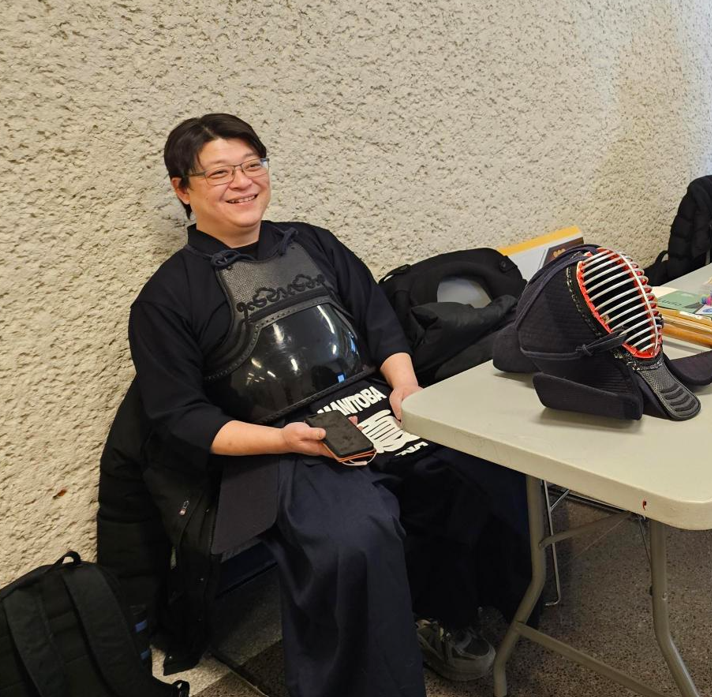 Person seated behind a table displaying kendo protective gear and equipment.