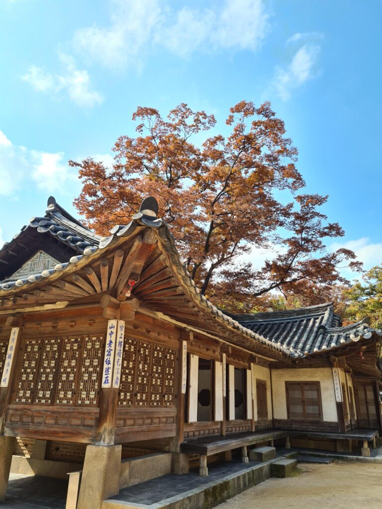 Traditional wooden temple building with carved details and a tiled roof, set beneath a tree with orange autumn leaves against a blue sky.