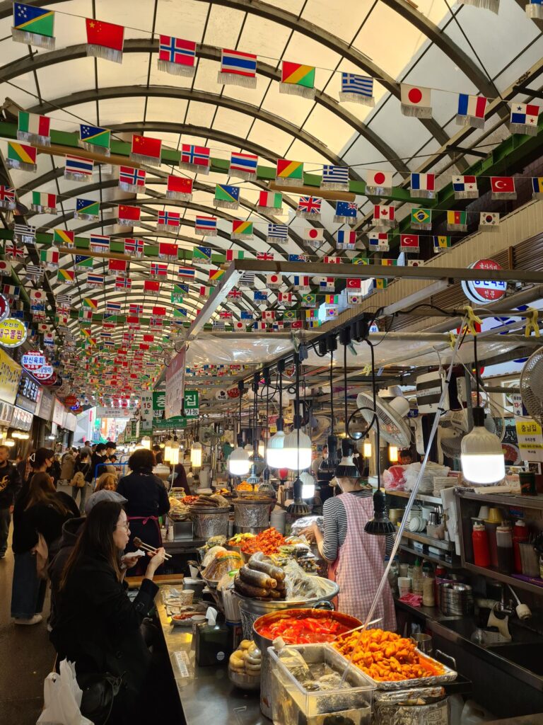 Busy indoor market aisle filled with food stalls, hanging lights, and rows of small international flags suspended from the ceiling.