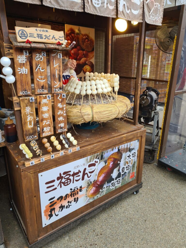 Small wooden street food stall displaying trays of round pastries and skewers, with handwritten signs and lanterns hanging above the counter.