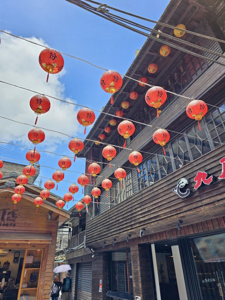 Traditional street lined with wooden buildings, where rows of red lanterns hang overhead against a bright blue sky.