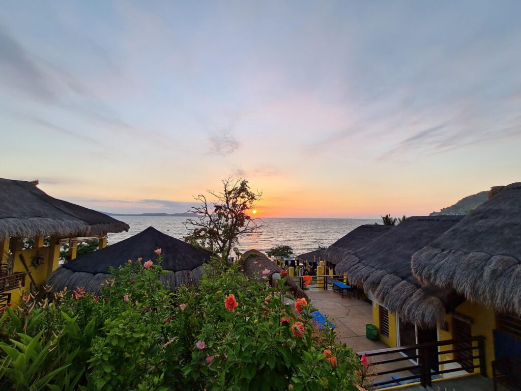 Sunset view over thatched-roof cabins and lush gardens, with the sun setting over the ocean and hills in the distance.