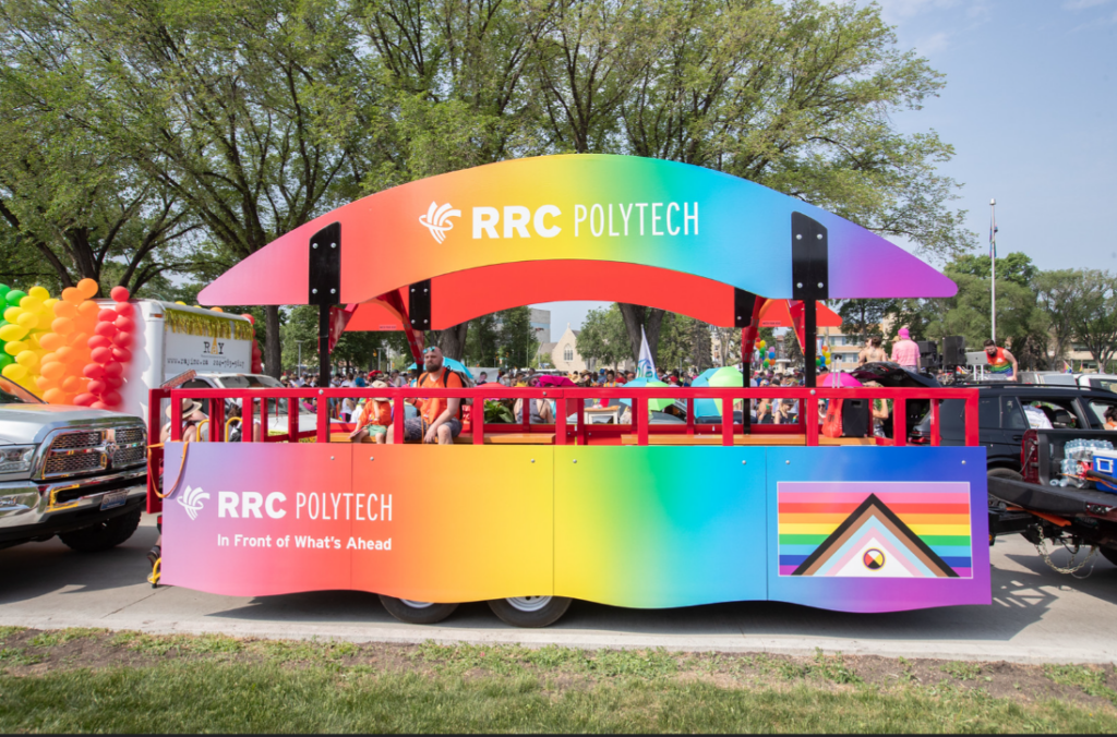 RRC Polytech Pride parade float with a rainbow-coloured canopy and panels, featuring the text “RRC Polytech – In Front of What’s Ahead,” driving along a tree-lined street with people seated inside and others gathered around.