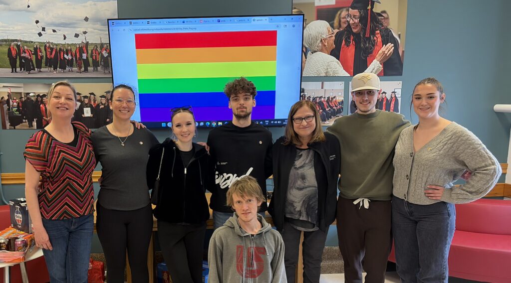 A group of eight people posing indoors in front of a screen displaying a rainbow Pride flag, with framed graduation photos on the wall behind them.