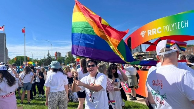Person waving a large rainbow Pride flag in a grassy area near an RRC Polytech parade float, with a crowd gathered for a Pride event.
