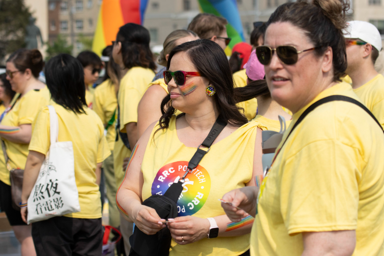 Participants in yellow RRC Polytech Pride t‑shirts stand together outdoors, including one person with a rainbow face stripe and Pride accessories, with a crowd gathered behind them.