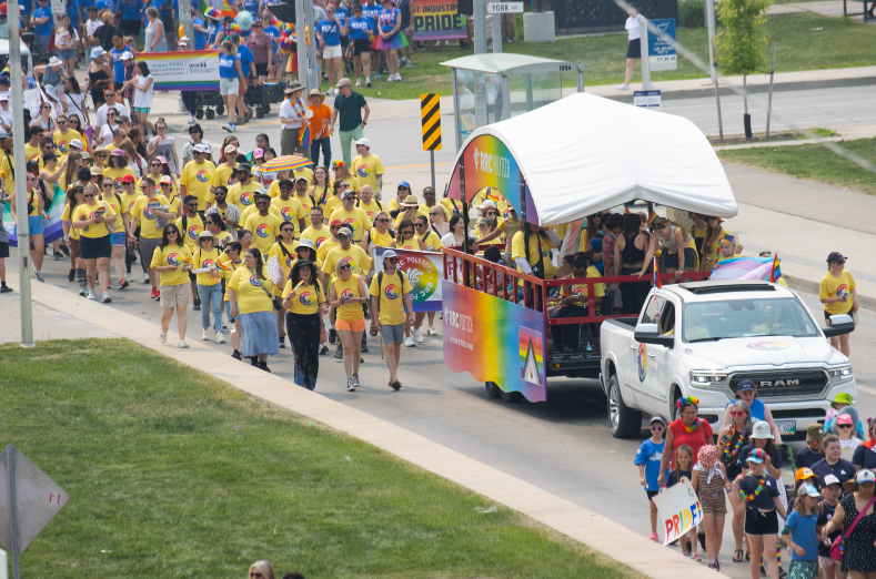 Wide view of a Pride parade showing a truck pulling an RRC Polytech rainbow float, surrounded by a large group of participants in yellow shirts walking along a city street.
