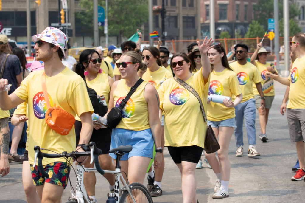 RRC Polytech participants in yellow Pride t-shirts walk together down a city street, some waving and carrying water bottles, with a bicycle and small Pride flags visible.