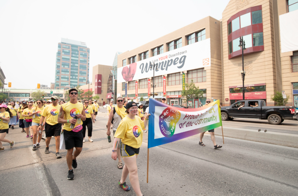 Large group of RRC Polytech participants walking downtown carrying a banner that reads “Proud to Be Part of the Community,” with buildings and spectators in the background.