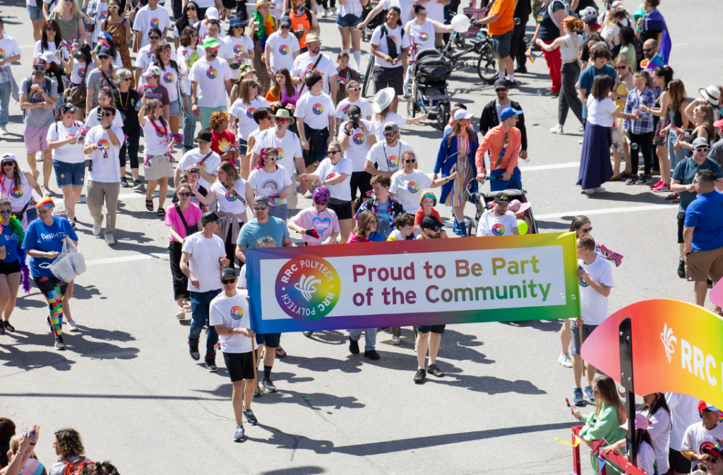 Large group of parade participants wearing white RRC Polytech shirts carry a rainbow banner that reads “Proud to Be Part of the Community,” with a crowded street scene around them.