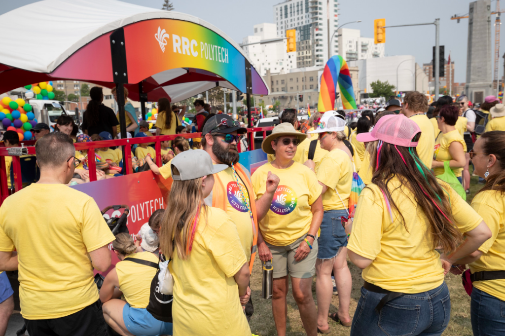 RRC Polytech participants in yellow Pride t-shirts gathered and talking beside a rainbow-coloured parade float in an urban setting.