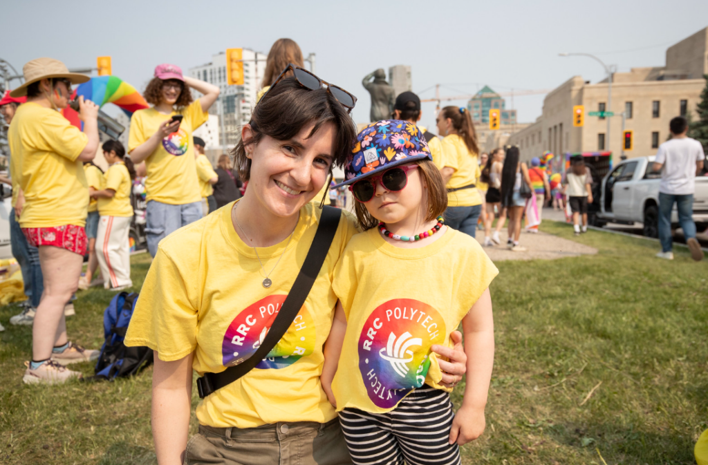 Adult and child in matching yellow RRC Polytech Pride shirts stand together in a grassy area during a Pride celebration, with a crowd, rainbow flags, and city streets behind them.