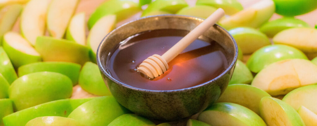 A bowl of honey sits in the middle of sliced green apples.