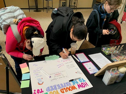 Community members participate in an International Pronouns Day activity by writing on a board titled “How Would You Like to Be Referred To?” and exploring pronoun resources at an information booth.
