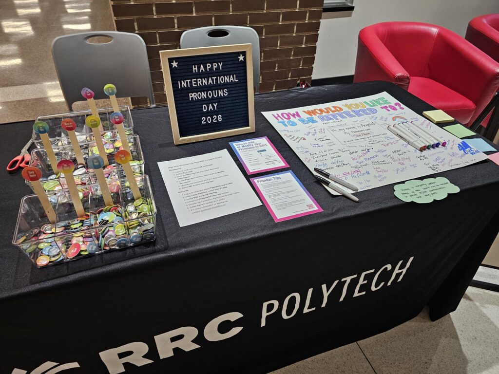 Table display for International Pronouns Day at RRC Polytech featuring pronoun pins, information cards, markers, sticky notes, a community participation board, and a sign reading “Happy International Pronouns Day 2026.”
