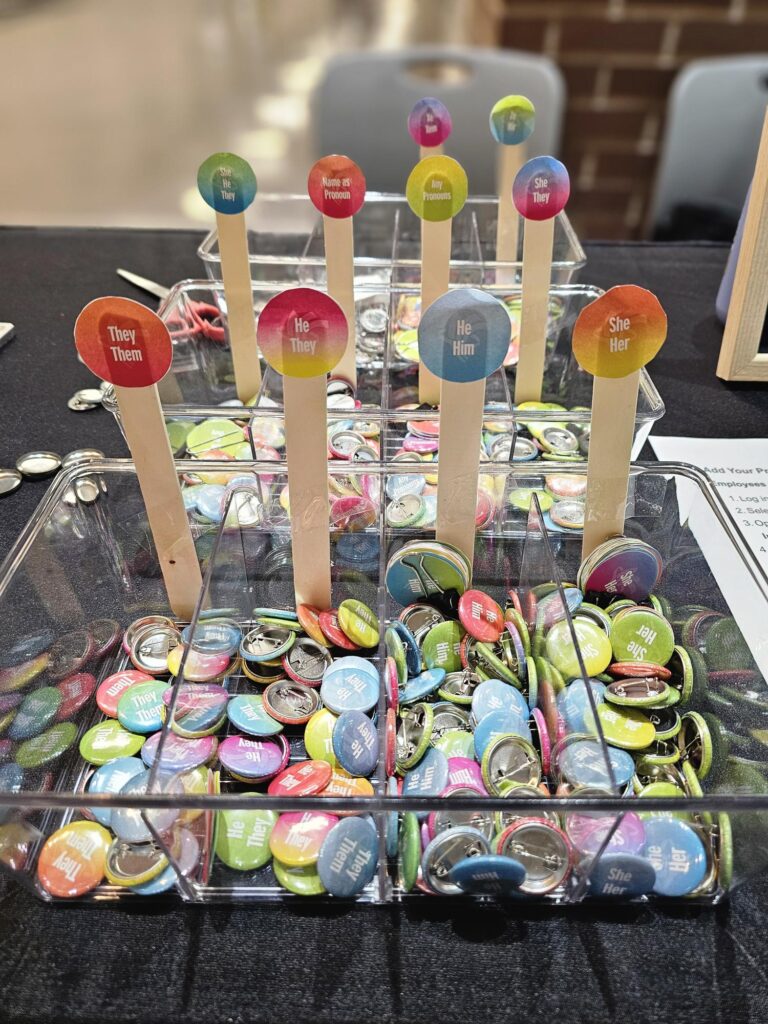 Close-up of a pronoun pin display with colourful buttons representing a variety of pronouns, including she/her, he/him, they/them, she/they, he/they, any pronouns, and name as pronoun.