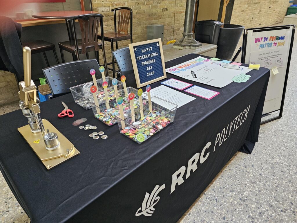 Table display for International Pronouns Day at RRC Polytech featuring pronoun pins, information cards, markers, sticky notes, a community participation board, and a sign reading “Happy International Pronouns Day 2026.”