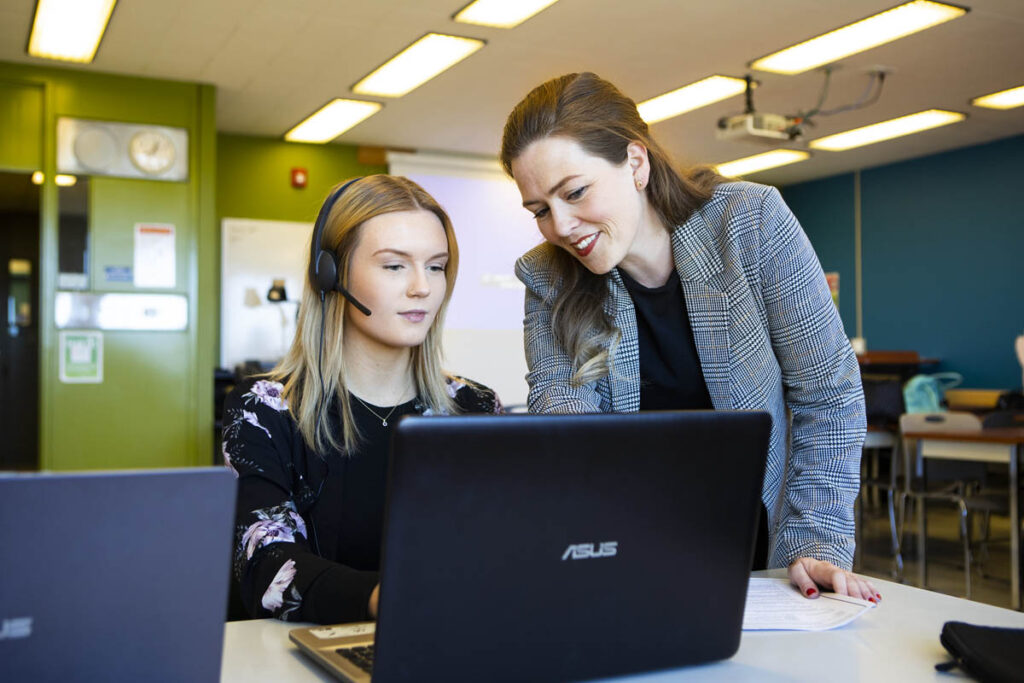 Instructor talking to a student wearing a headset and working on a laptop in a classroom