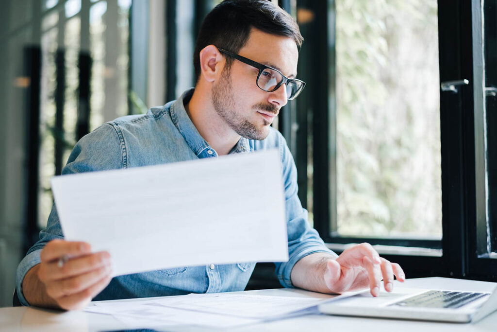 Man holding a sheet of paper and working on a laptop