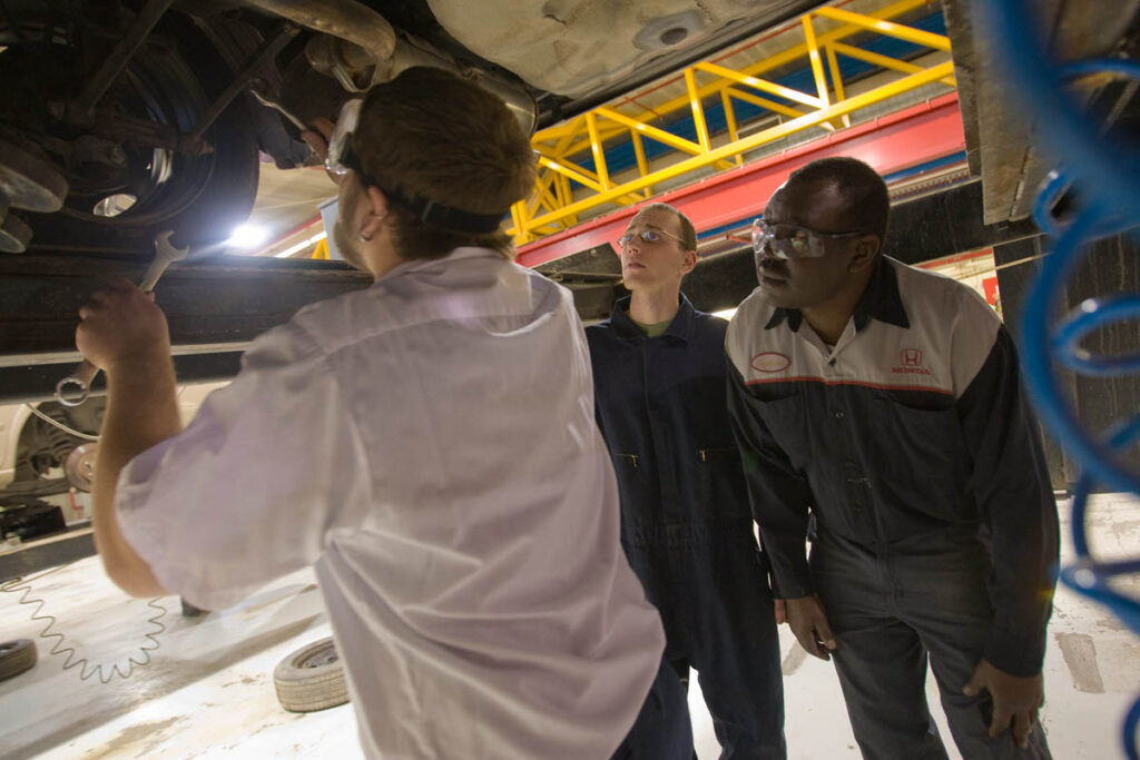 Automotive students working underneath a raised up vehicle in a shop while an instructor watches
