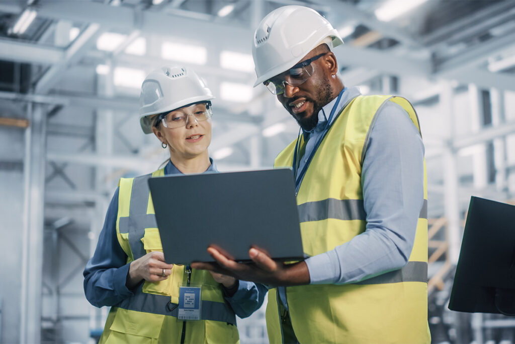 Two people in a warehouse wearing yellow safety vests and looking at a laptop