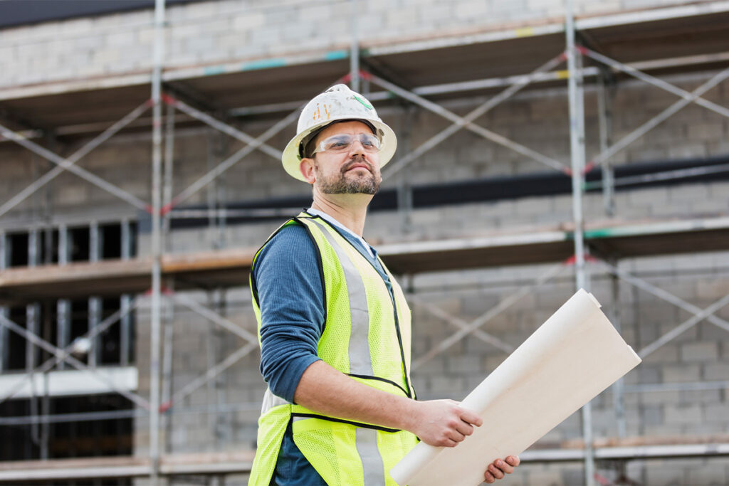 Construction worker wearing yellow safety vest holding blueprints and staring off at job site