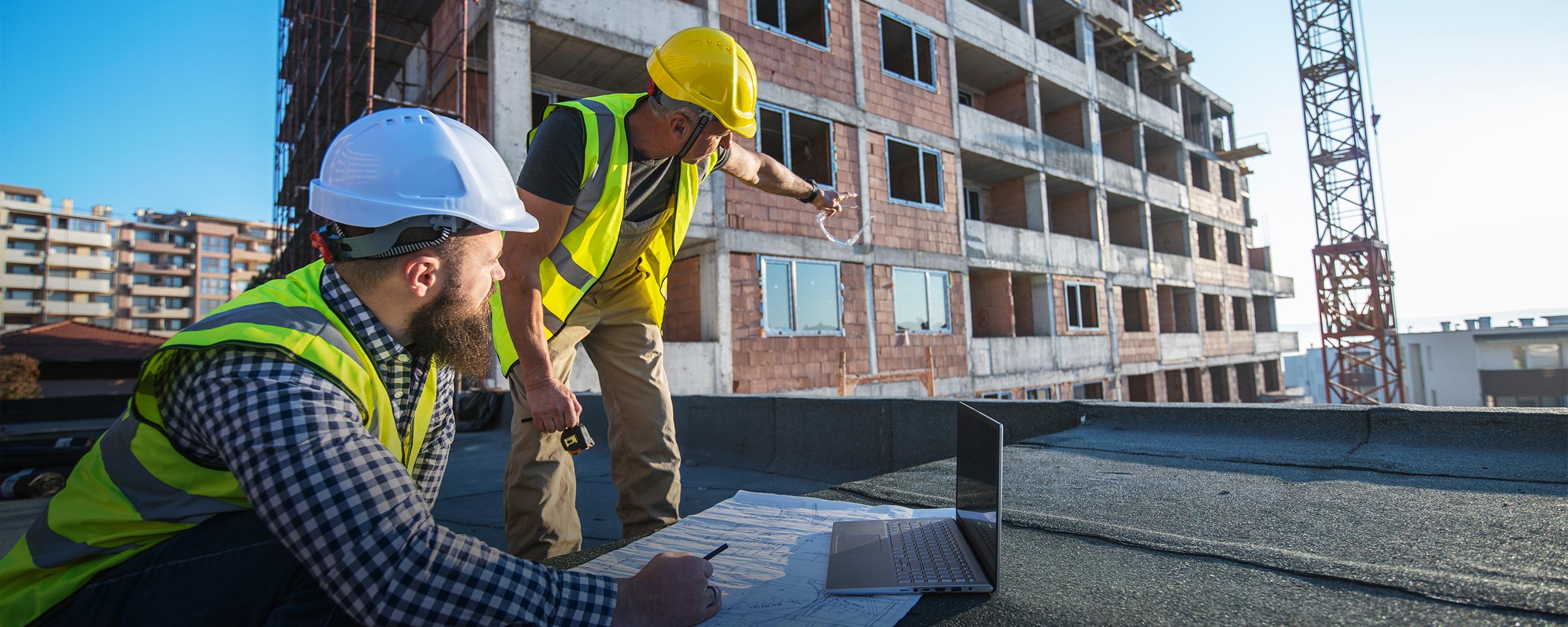 Two construction workers looking at blueprints and a laptop while on a construction site