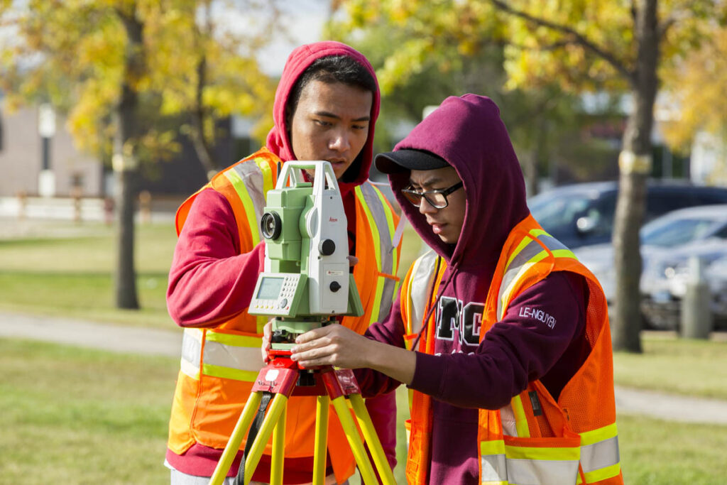 Two students wearing safety vests working with surveying equipment outdoors