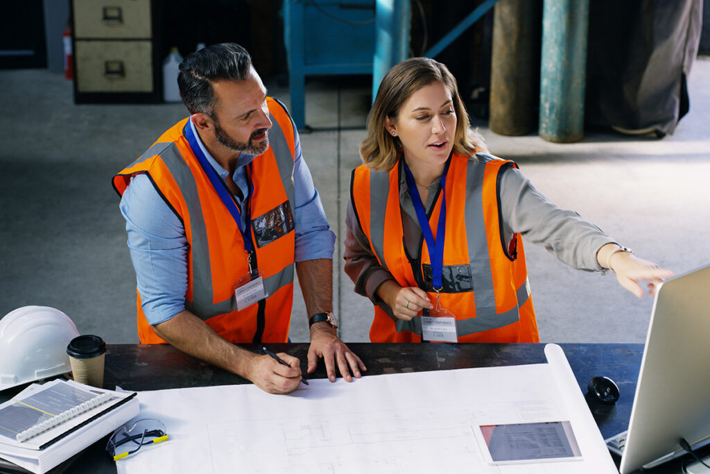 Two people wearing orange safety vests in a warehouse and pointing at something on a computer screen