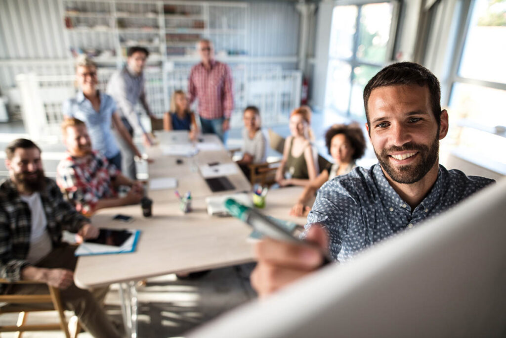 Man writing on a flip chart while a room full of colleagues watches on