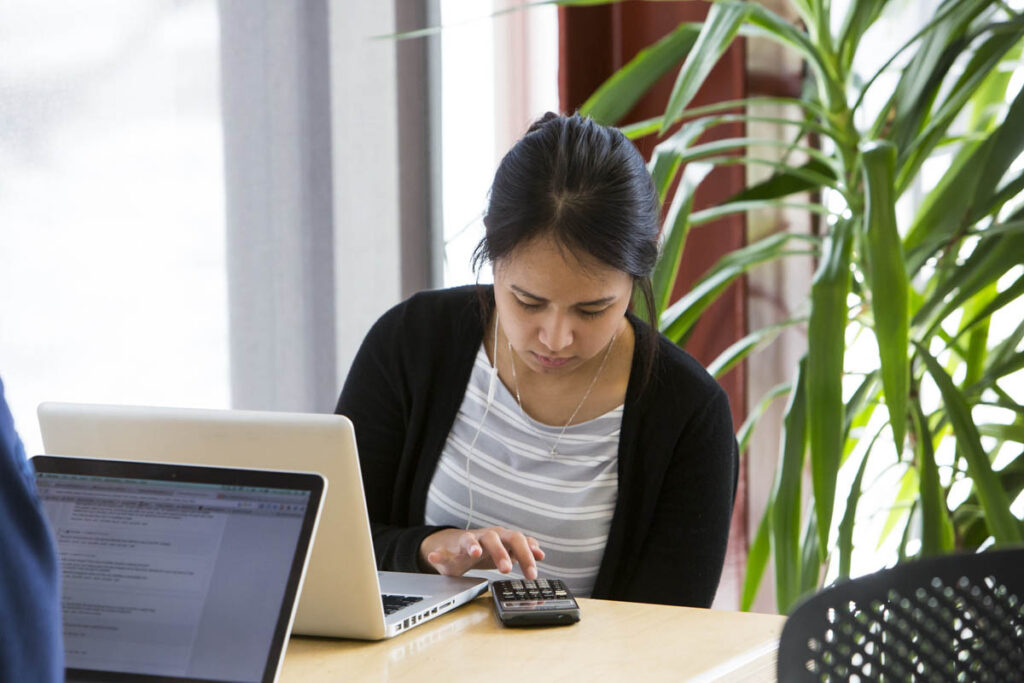 Woman sitting at a table with a laptop while typing on a calculator