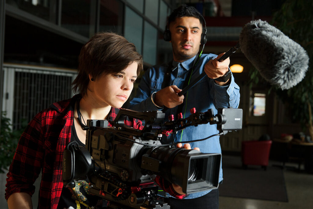 Two people on a film set, one holding a boom microphone and the other operating a camera